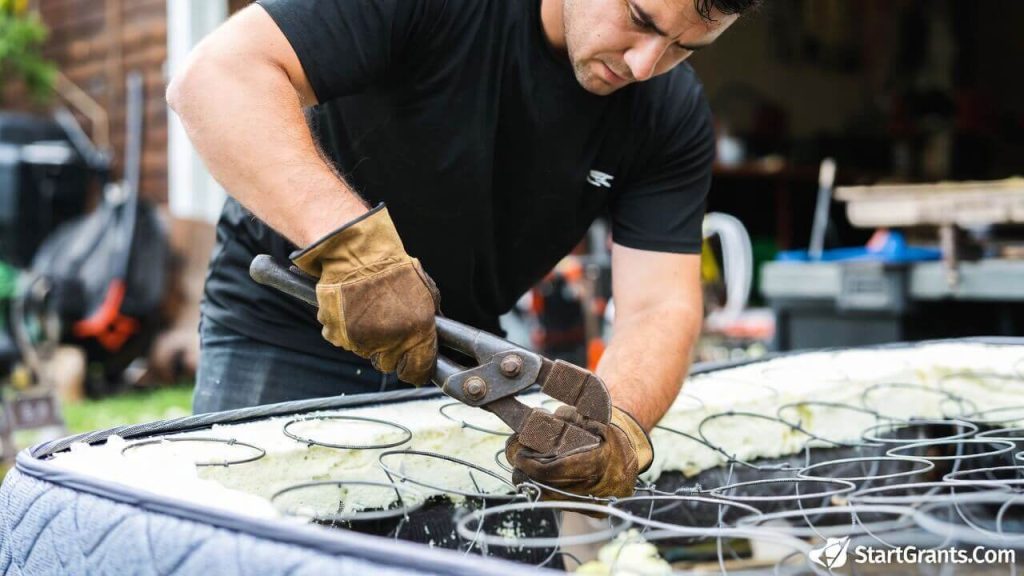A person wearing heavy work gloves using bolt cutters to dismantle an old mattress for scrap metal recycling.