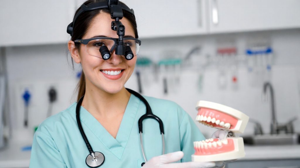 A smiling dental hygiene student wearing scrubs and dental loupes holding a typodont model.