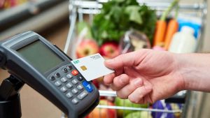 A woman's hand holding an EBT SNAP card at a grocery store card reader, with a cart full of fresh fruits and vegetables.