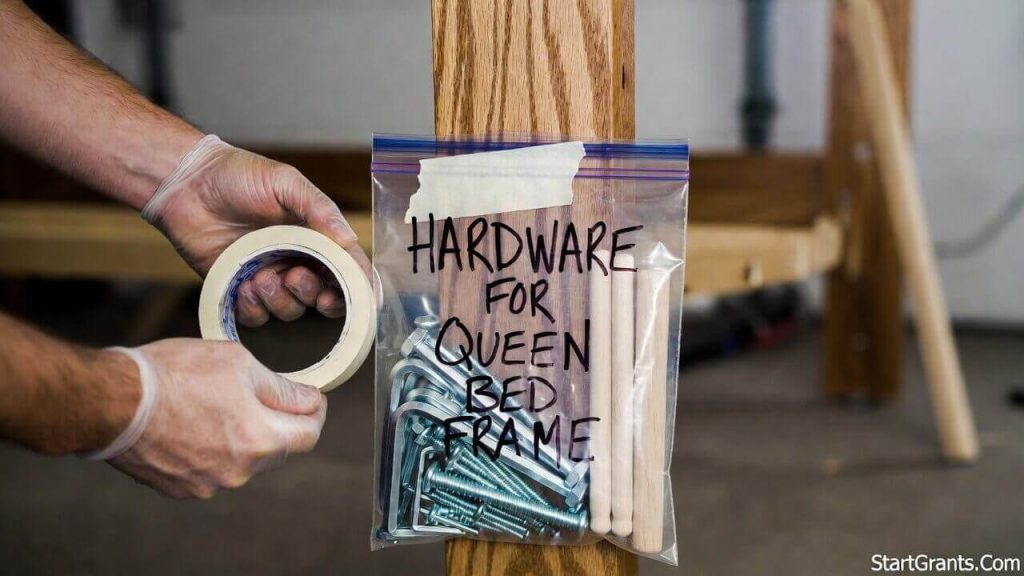 A close-up shot of hands using blue painter's tape to attach a Ziploc bag full of screws and bolts to a disassembled wooden bed frame leg.