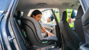 A smiling mother successfully installing a brand new convertible car seat in the back seat of a car, with a certified safety technician watching.