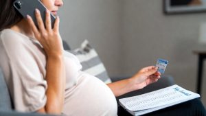 Pregnant woman holding a smartphone and an insurance card, calling her Medicaid provider for prenatal rewards.