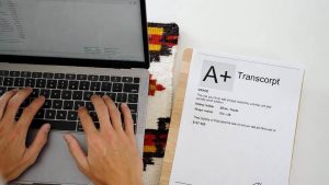 A student typing on a laptop with an official academic transcript on the desk