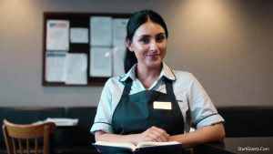 A smiling worker in a coffee shop apron (like Starbucks) holding a college textbook during a break.