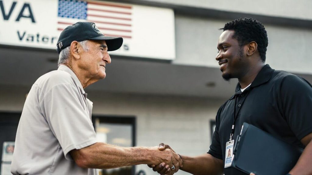 A military veteran shaking hands with a housing support worker outside a VA-affiliated facility.