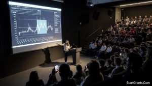 A female graduate student presenting her research data on a large screen at a national academic conference symposium.