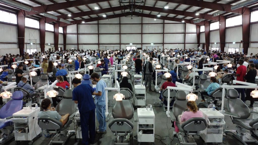 A large gymnasium filled with portable dental chairs providing free care to hundreds of people.