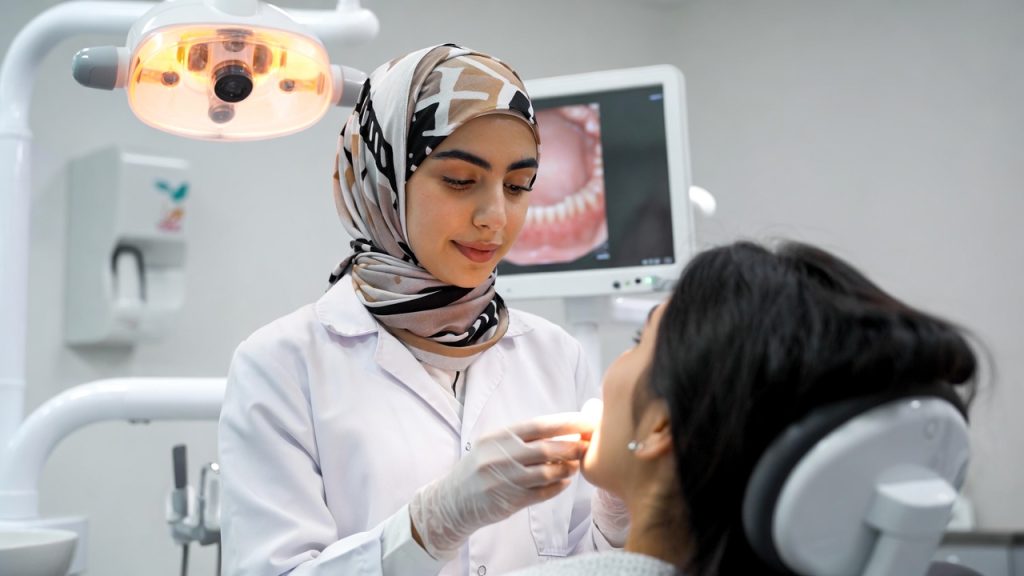 A Muslim dental hygiene student wearing a hijab and lab coat interacting with a patient.