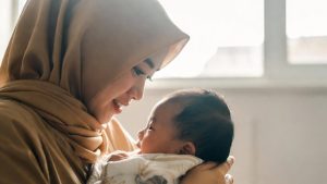 Seorang ibu tunggal yang tersenyum sedang bekerja di laptop di ruang tamu yang rapi, sementara anaknya yang masih balita bermain dengan tenang di dekatnya.