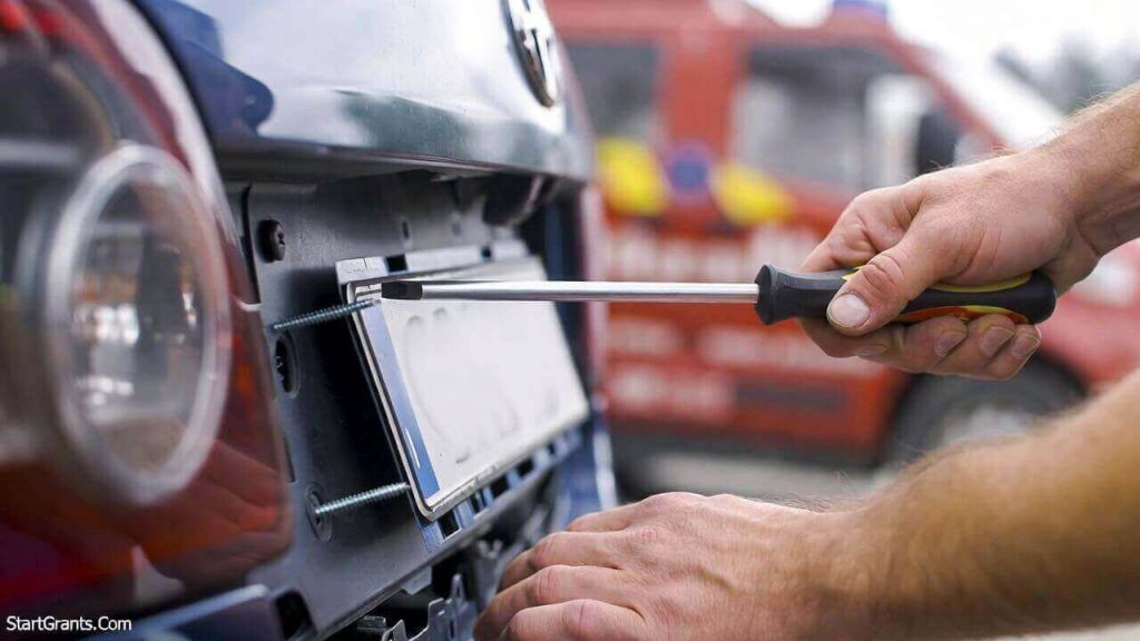 A car owner removing their license plates with a screwdriver before handing the vehicle over to a charity tow truck.